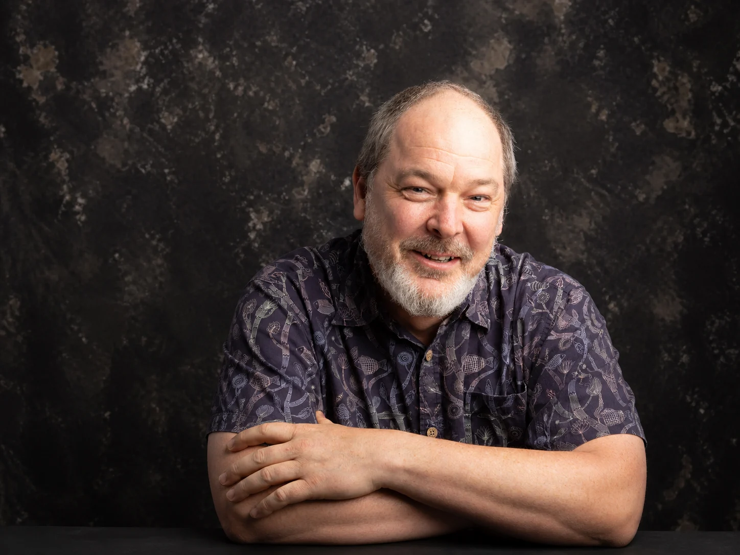 Portrait of Dylan Sisson wearing glasses and a patterned dark shirt against a dark studio background.