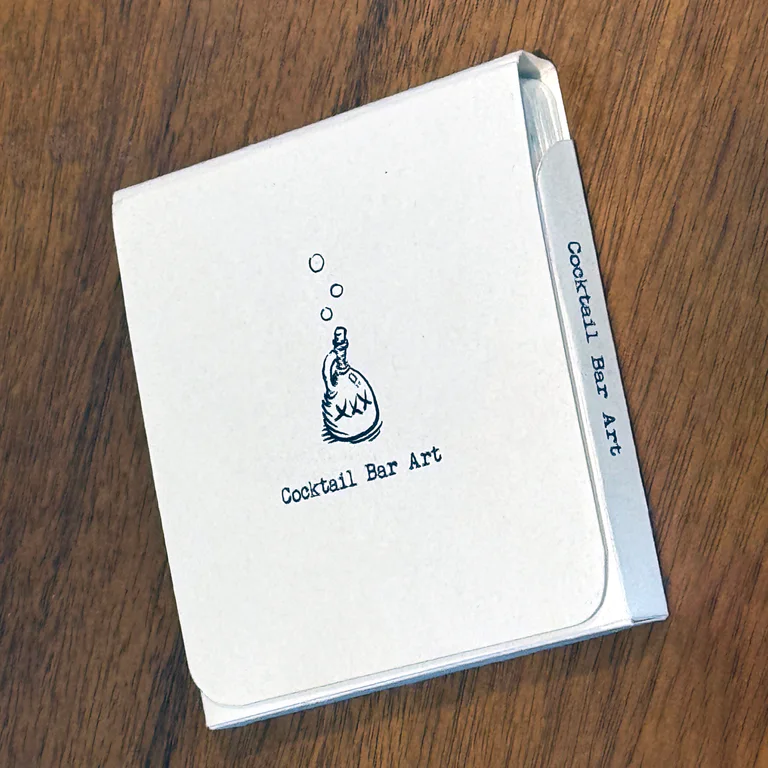 A handmade white folio labeled Cocktail Bar Art resting on a wooden table.