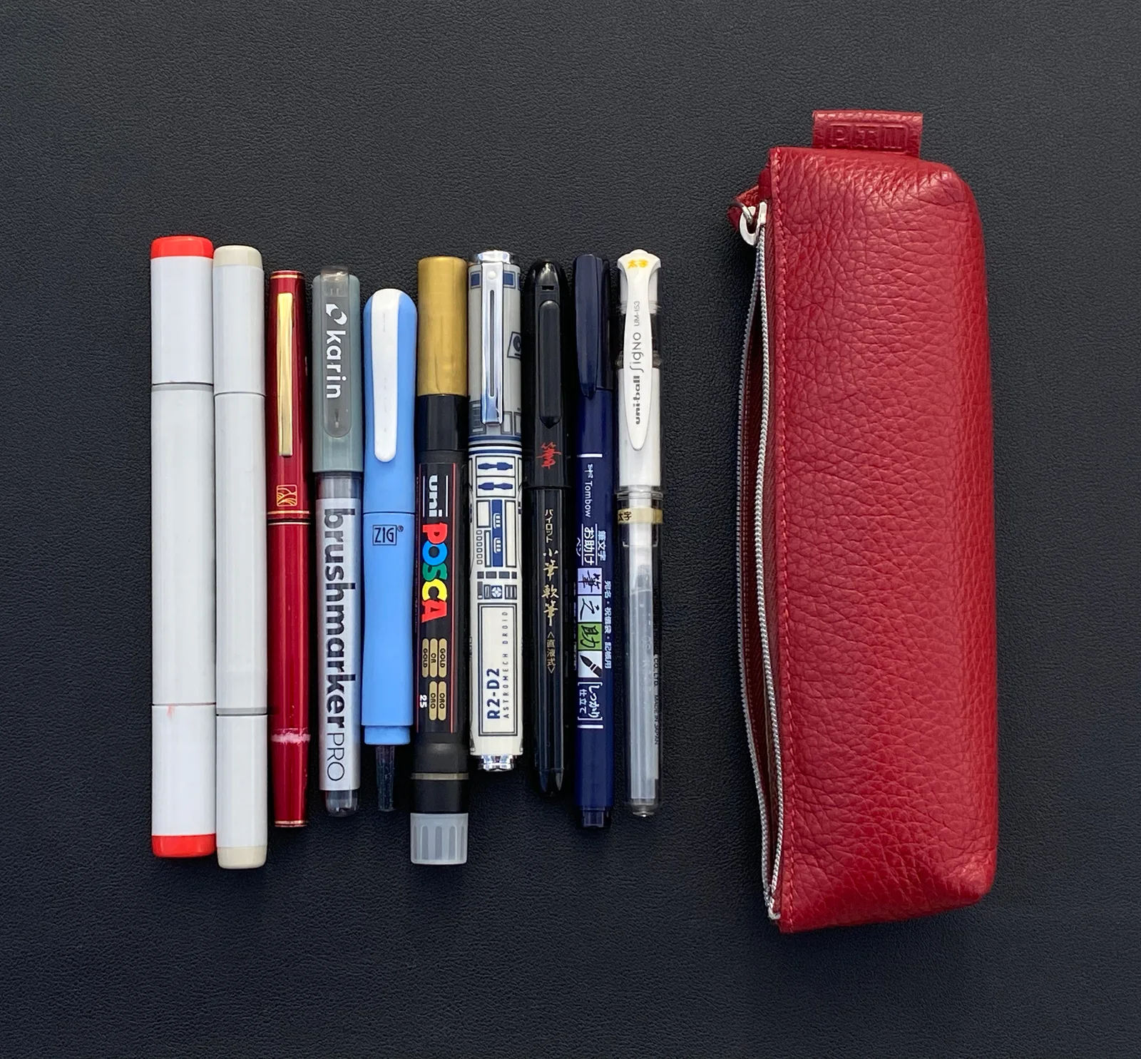 An assortment of brush pens, gel pens, markers, and drawing tools arranged beside a red leather pencil case.