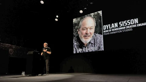 Dylan Sisson speaking onstage beside a projected portrait and his Pixar RenderMan role.